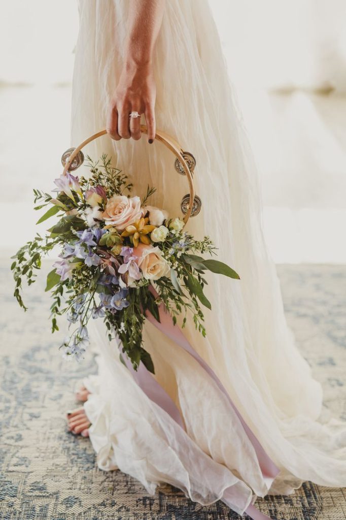 In questa foto una sposa con abito scivolato stile boho chic tiene nella mano sinistra un hoop bouquet a tamburello con rose bianche, fiori lilla, foglie e un nastro lungo colore lilla