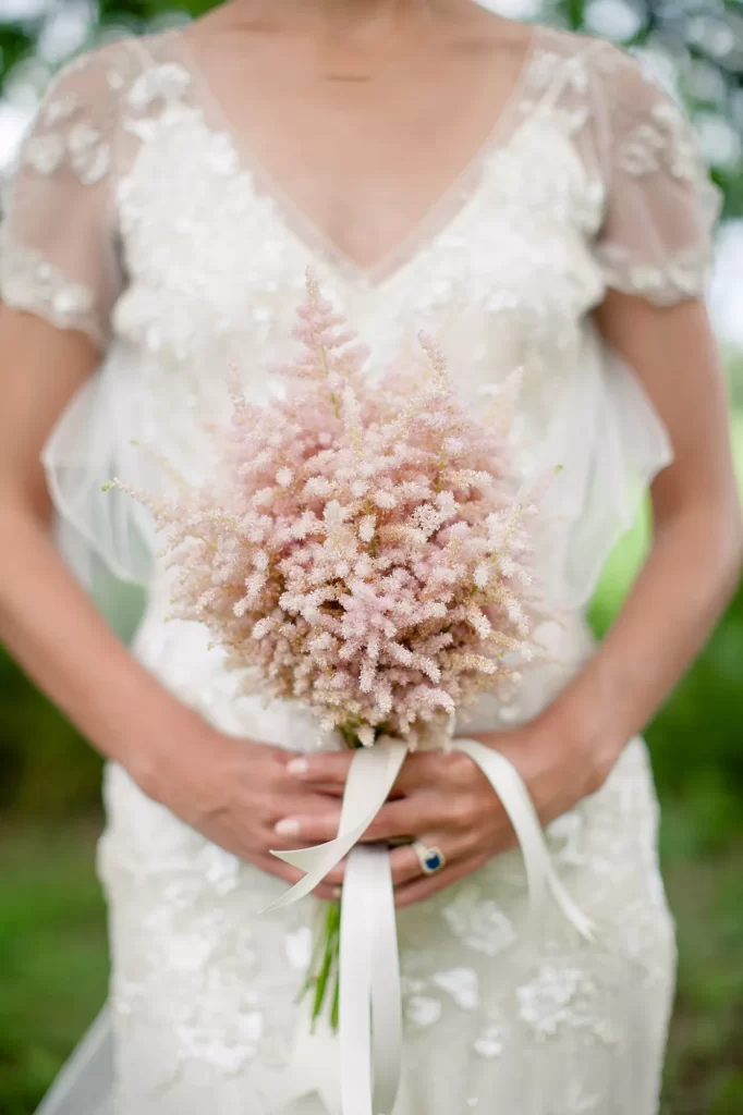 In questa foto le mani di una sposa che tengono un piccolo bouquet di astilbe rosa legate da un nastro bianco. La sposa indossa un abito di pizzo con manichette e scollo a v