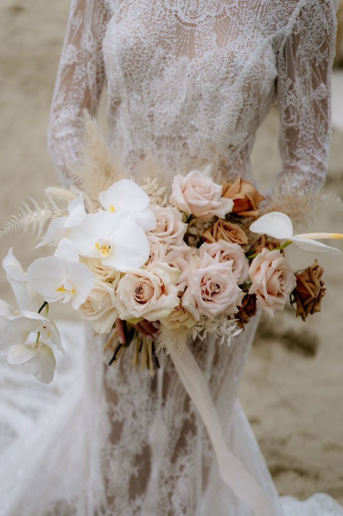 In questa foto una sposa con abito a sirena in pizzo macramè tiene tra le mani un bouquet con rose rosa, rose cappuccino, orchidee bianche, felce bianca tutte legate da un lungo nastro di lino bianco