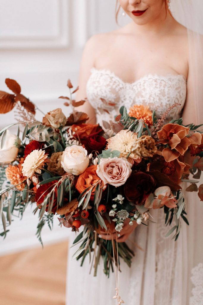 In questa foto una sposa tiene tra le mani il suo bouquet autunnale realizzato con rose cappuccino, gerbere rosa, rose rosse, zinnia arancione, foglie d'edera e rami di ulivo