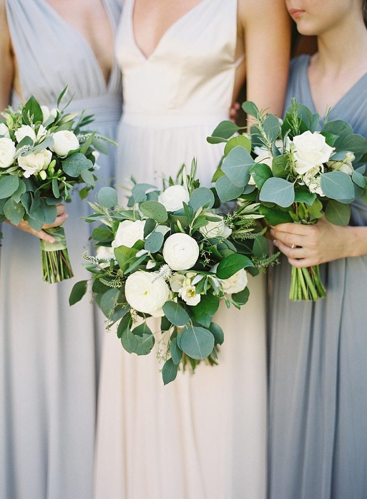 In questa foto le mani di una sposa e di due damigelle che tengono tre bouquet di fiori bianchi ed eucalipto. Le damigelle indossa un abito con scollo all'americana colore carta da zucchero