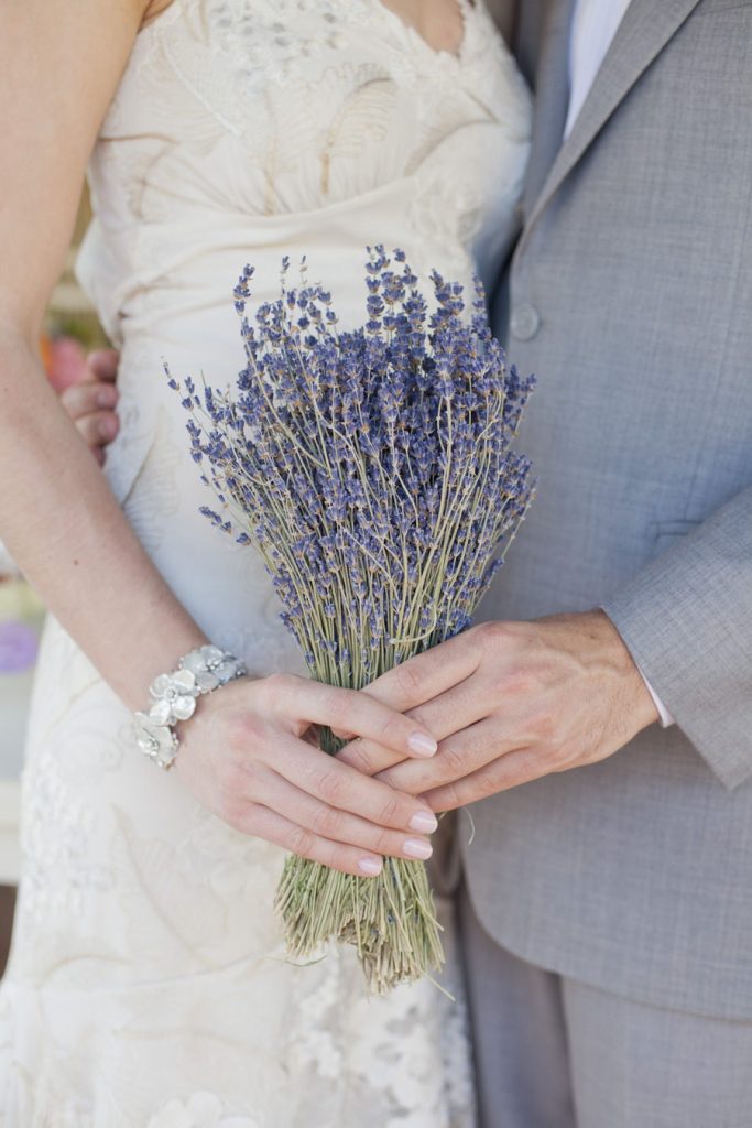 In questa foto le mani di due sposi che tengono un bouquet di lavanda. La sposa indossa un bracciale di madreperla, mentre lo sposo indossa un abito grigio perla