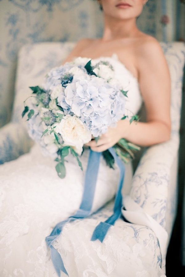 In questa foto una sposa seduta su una poltrona bianca damascata tiene nella mano sinistra un bouquet di ortensie celesti e rose inglesi legate da un nastro di velluto avion