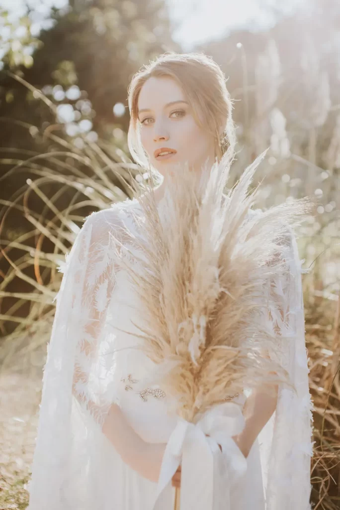 In questa foto una sposa in un giardino tiene tra le mani un bouquet di pampas legate da un nastro bianco di seta