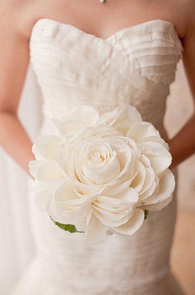 In questa foto le mani di una sposa che tengono un bouquet composite con un solo fiore grande bianco a forma di rosa