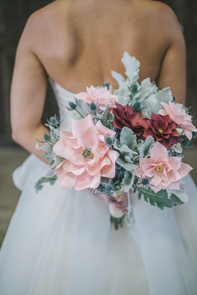In questa foto una sposa di spalle mentre tiene tra le mani un bouquet di foglie e fiori di carta rosa e rossi