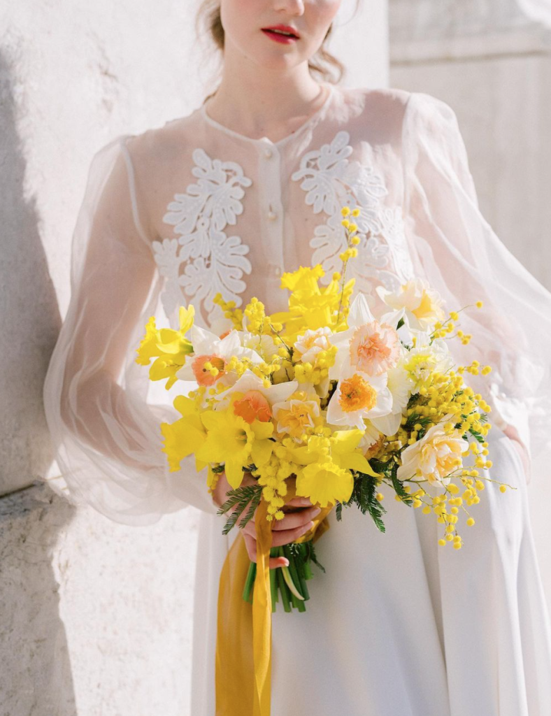 In questa foto una sposa con abito scivolato e maniche balloon di seta trasparente con ricami sul petto tiene nella mano destra un bouquet estivo con fiori gialli e bianchi