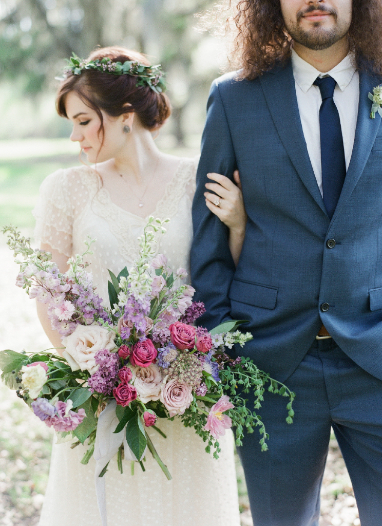 In questa foto una sposa si tiene con il braccio sinistro al braccio destro del suo sposo e nella mano destra tiene un bouquet scomposto di rose fucsia,  bianche e lilla e delphinium lilla e bianco