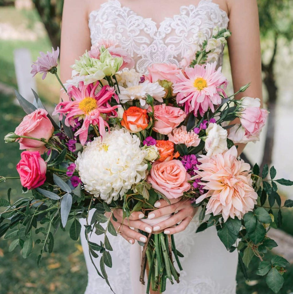 In questa foto una sposa tiene tra le mani un bouquet scomposto di rose rosa e arancioni, dalie rosa e bianche e gerbere rosa e lilla