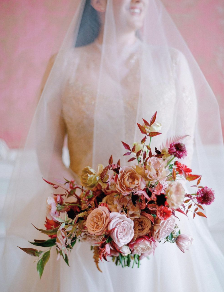 In questa foto una sposa tiene tra le mani un bouquet scomposto di rose e fiori di campo nei toni del rosa e del fucsia. La sposa indossa un velo con calata