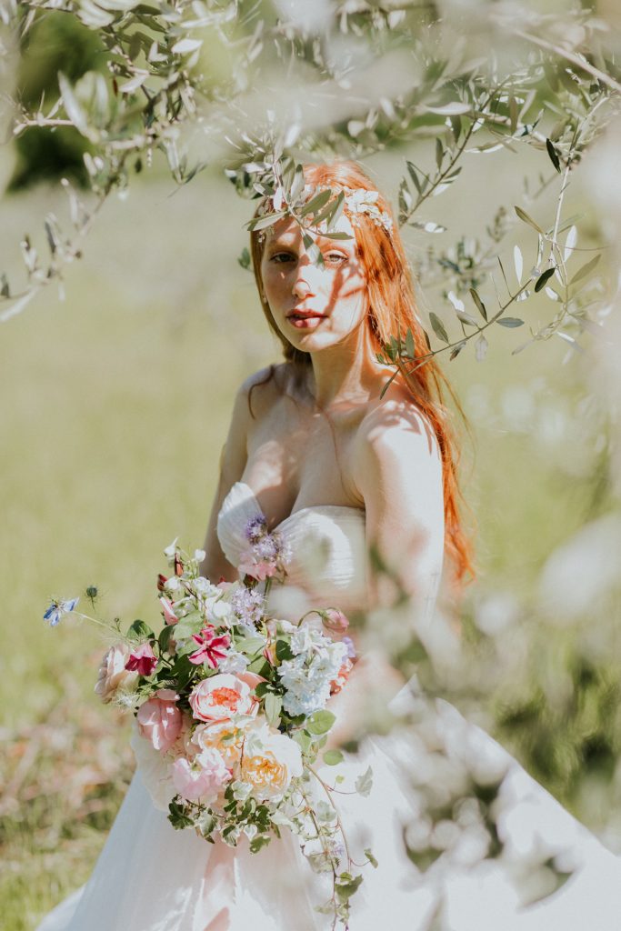 In questa foto una sposa con i capelli rossi posa tra gli ulivi mentre tiene tra le mani un bouquet scomposto  di rose inglesi colore pesca, gialle, rosa e fiori di campo fucsia e lilla