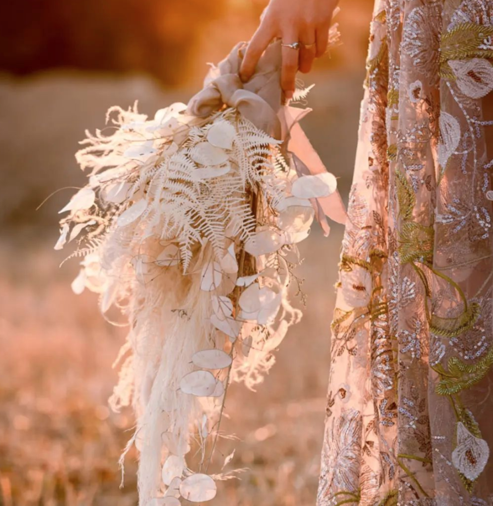 In questa foto una sposa tiene nella mano destra un bouquet nei toni del bianco e del beige con felci, pampas e petali stretti da un nastro colore rosa cipria