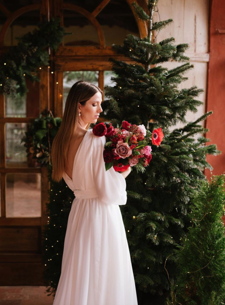 In questa foto una sposa con capelli sciolti e lisci e abito scivolato scollato sulla schiena posa davanti ad un albero di Natale, tenendo e guardando il suo bouquet invernale con fiori rossi