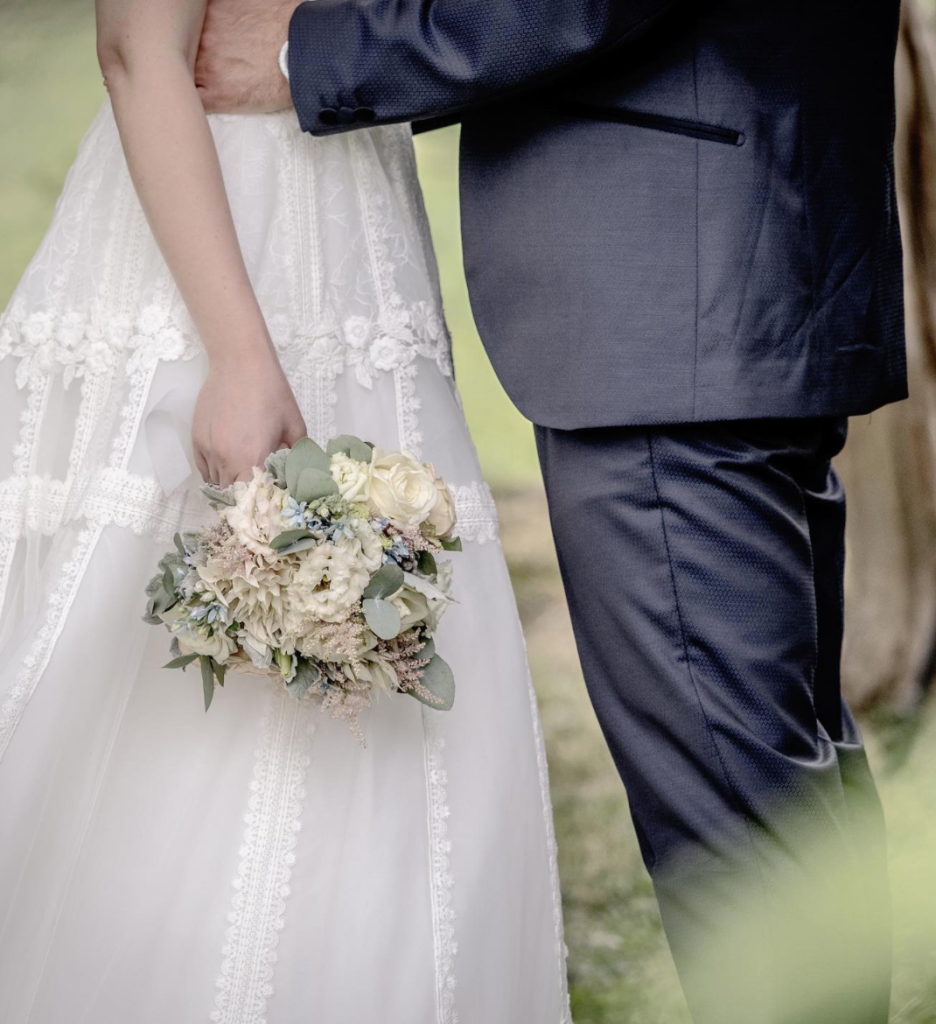 In questa foto una sposa abbracciata dallo sposo tiene nella mano destra un mazzolino di fiori bianchi ed eucalipto