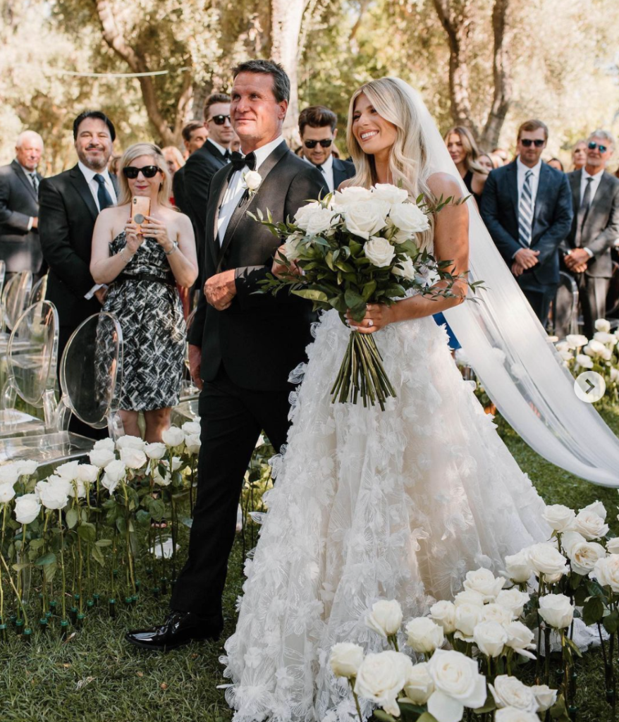 In questa foto una sposa attraversa al braccio del papà un corridoio di rose bianche su un prato circondata dagli ospiti tenendo in mano un grande bouquet di rose bianche e felci. La sposa indossa un abito a trapezio con fiori 3D sulla lunghezza e velo lungo a taglio vivo