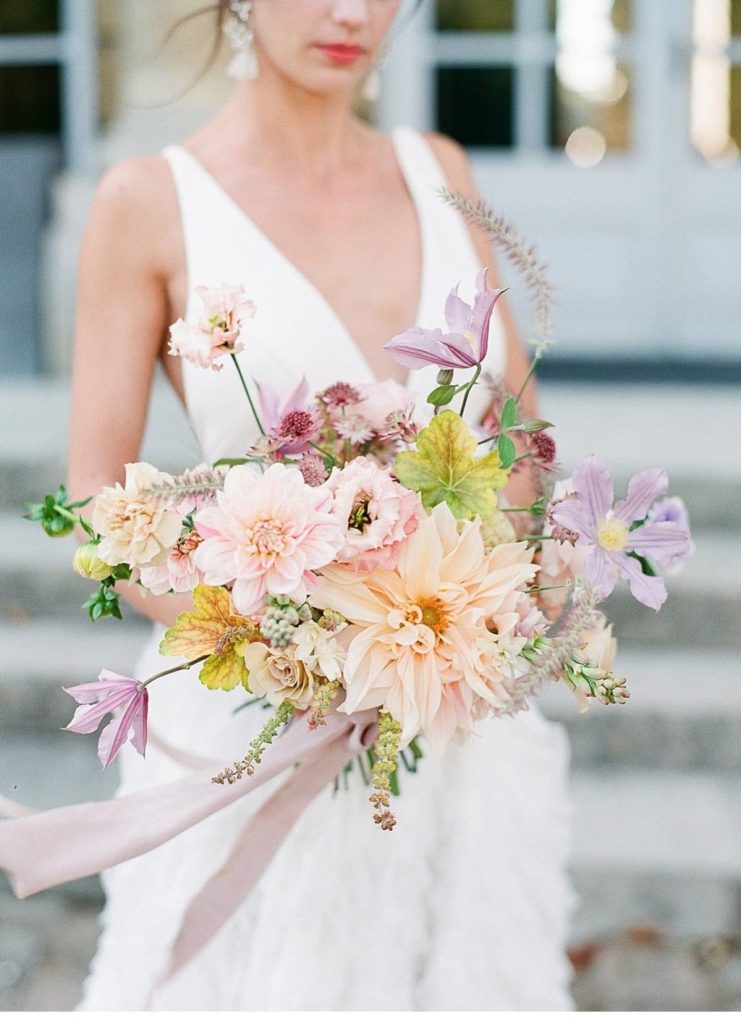 In questa foto una sposa con abito scivolato con scollatura a tiene nella mano destra un bouquet da sposa con dalie arancioni e rosa, fiori lilla e gialli e foglie