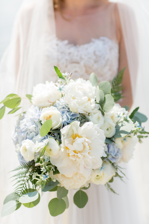 In questa foto le mani di una sposa che tiene un bouquet rotondo con dalie, peonie, rose e ranuncoli bianchi, ortensie celesti, eucalipto e felce