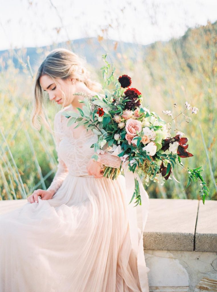 In questa foto una sposa con abito scivolato colore cipria seduta su un muretto immerso in un prato tiene nella mano sinistra un bouquet scomposto con rose rosa, fiori bordeaux e foglie di eucalipto