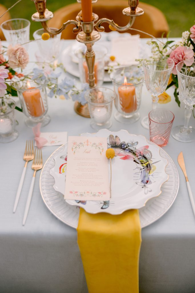 In questa foto una mise en place elegante per un ricevimento sul prato nei toni del giallo e del rosa con un menù di matrimonio rettangolare poggiato su un piatto decorata con farfalle dipinte. Sotto il piatto è disteso un tovagliolo giallo pendente lungo la tovaglia