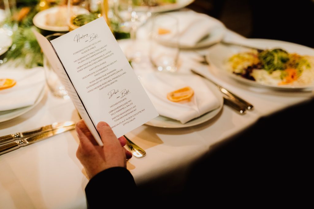 In questa foto l'invitato ad un ricevimento tiene nella mano sinistra un menù matrimonio a libro e legge le portate. Dietro è presente una tavola imbandita 