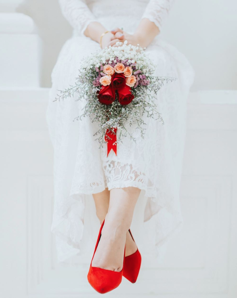 In questa foto le gambe di una sposa seduta a penzoloni che indossa décolleté rosse e tiene poggiato sulle ginocchia un bouquet di rose rossa e rosa, nebbiolina e piccoli fiori lilla