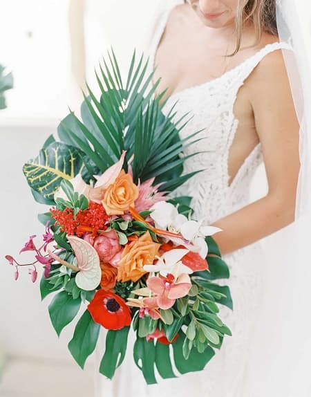 In questa foto, una sposa tiene in mano un bouquet creato con fiori stile matrimonio tropicale dai toni accesi e foglie di grandi dimensioni 