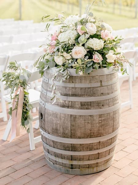In questa foto, una composizione di ortensie e rose bianche e rosa, arricchita da rami di ulivo ed eucalipto, è sistemata sopra una botte di legno. Un esempio di fiori matrimonio stile Country