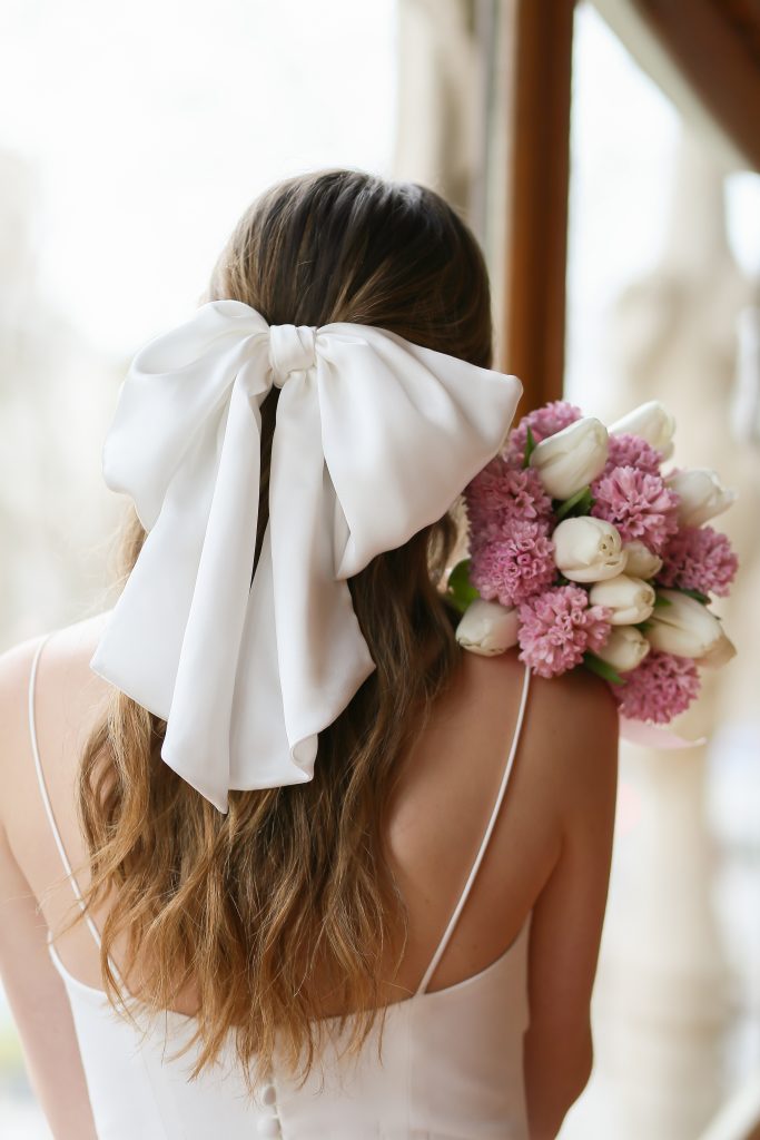 In questa foto una sposa di spalle con un grande fiocco bianco, tra gli accessori da sposa per capelli più originali di Blunbow.  Mostra anche il suo bouquet di tulipani bianchi e fiori colore rosa