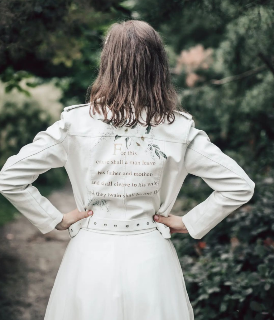 In questa foto una sposa con capelli corti di spalle con le mani sui fianchi mostra il retro del suo giubbotto di pelle bianco personalizzato con una citazione in inglese