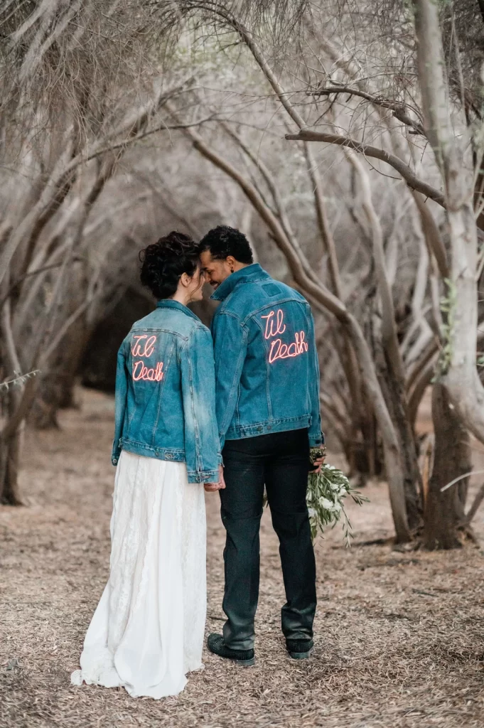 In questa foto due sposi in un bosco si tengono per mano e, l'uno appoggiato all'altra sulla fronte, mostrano il retro delle loro giacche jeans over personalizzate