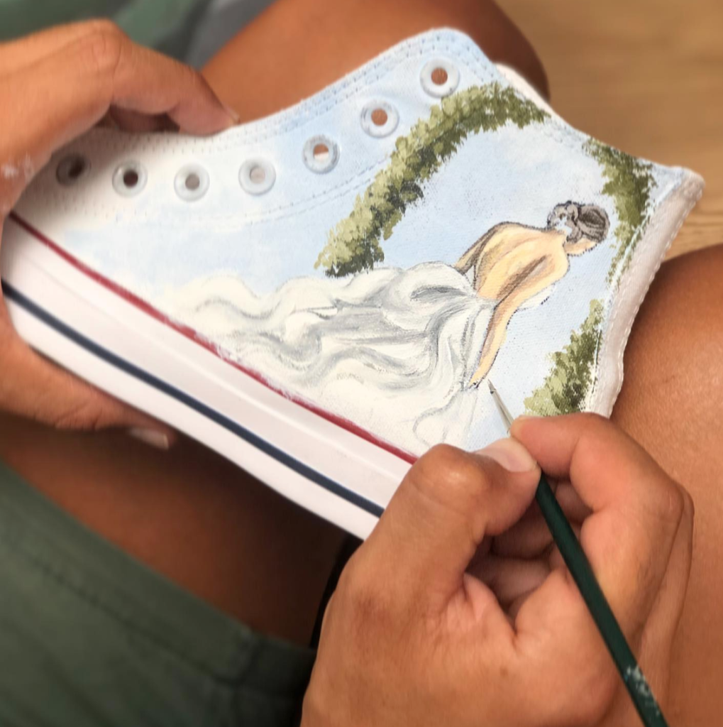 In questa foto la mano di un artista mentre dipinge una converse da sposa con il disegno di una sposa di spalle che varca un arco di di fiori