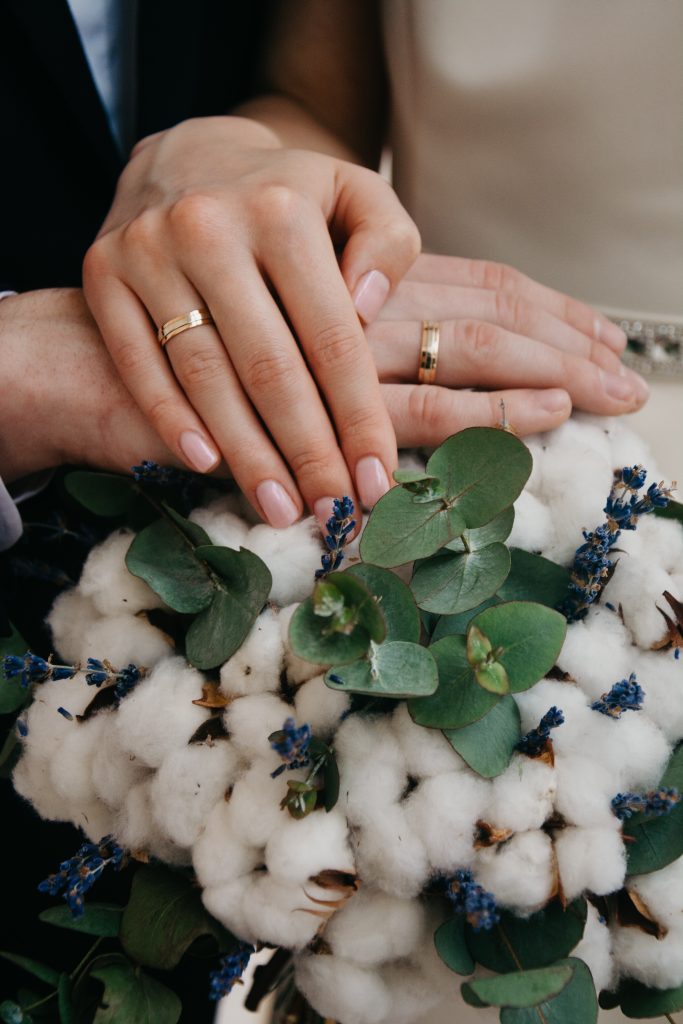 In questa foto le mani di due sposi una sopra all'altra su un bouquet di fiori di cotone mentre mostrano le loro fedi nuziali