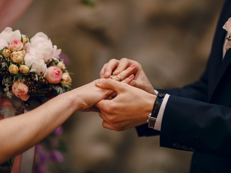 In questa foto le mani di due sposi mentre lo sposo mette al dito della sposa la fede nuziale. Lei porta nella mano destra un bouquet di rose colore rosa