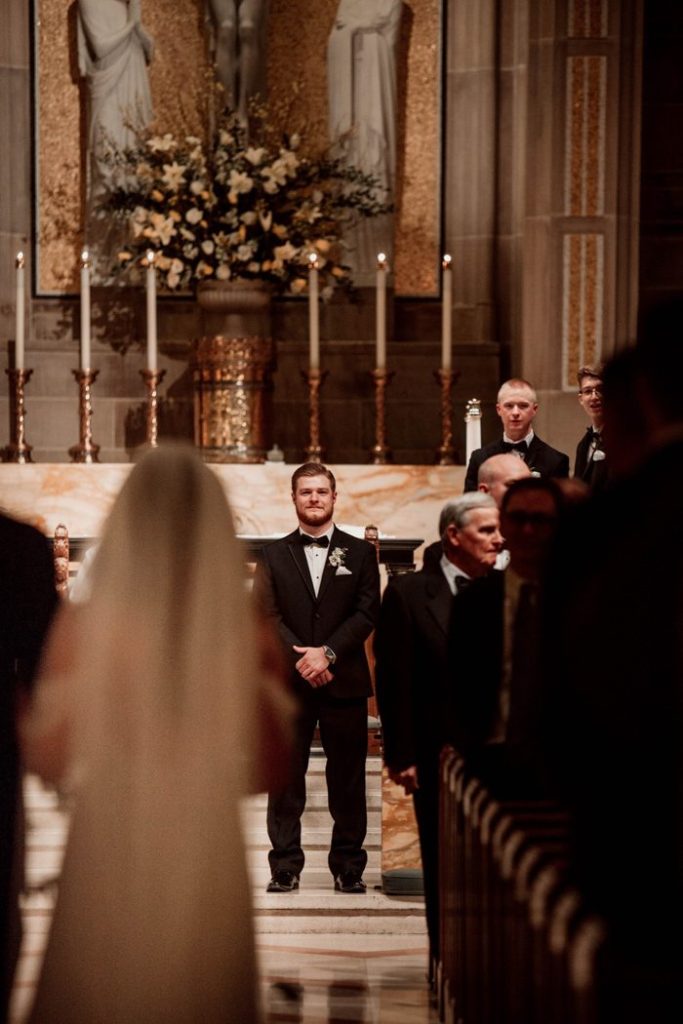 In questa foto uno sposo all'altare guarda l'ingresso della sposa in chiesa. Lo sposo è a fuoco mentre la sposa di spalle è sulla sinistra sfocata