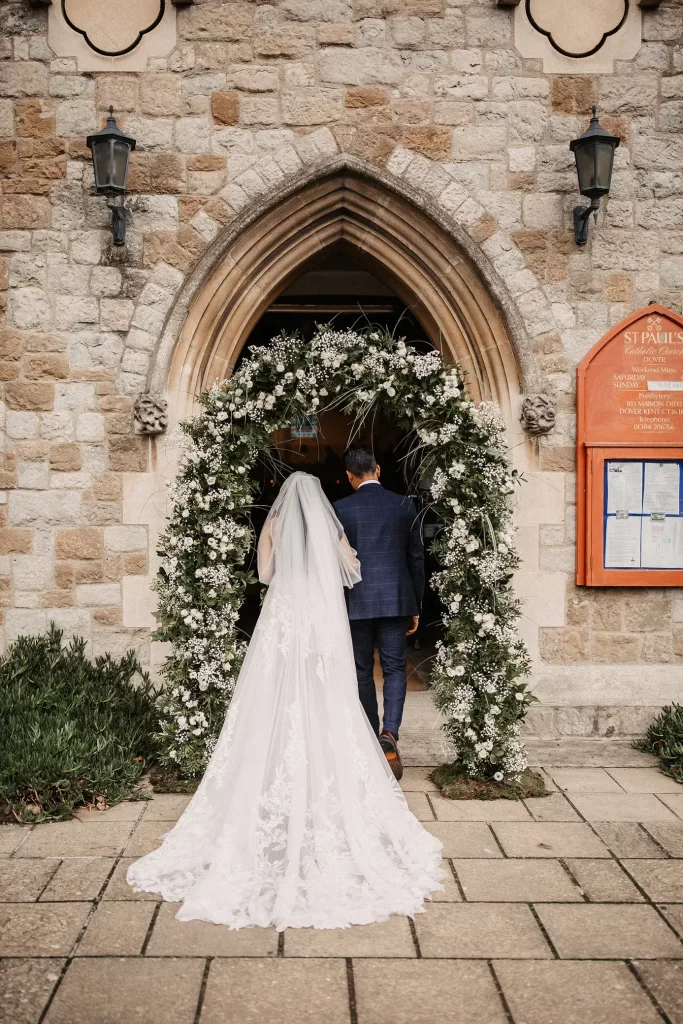 In questa foto l'ingresso della sposa al braccio del papà sulla soglia di una chiesa per matrimonio allestita da un arco di fiori bianchi
