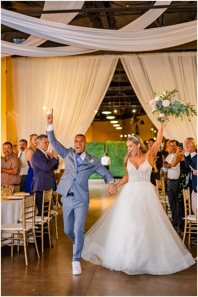 In questa foto l'ingresso sposi in sala ballando. Lo sposo indossa un abito colore carta da zucchero. La sposa un abito ad A in tulle e solleva in alto il suo bouquet di rose colore rosa ed eucalipto