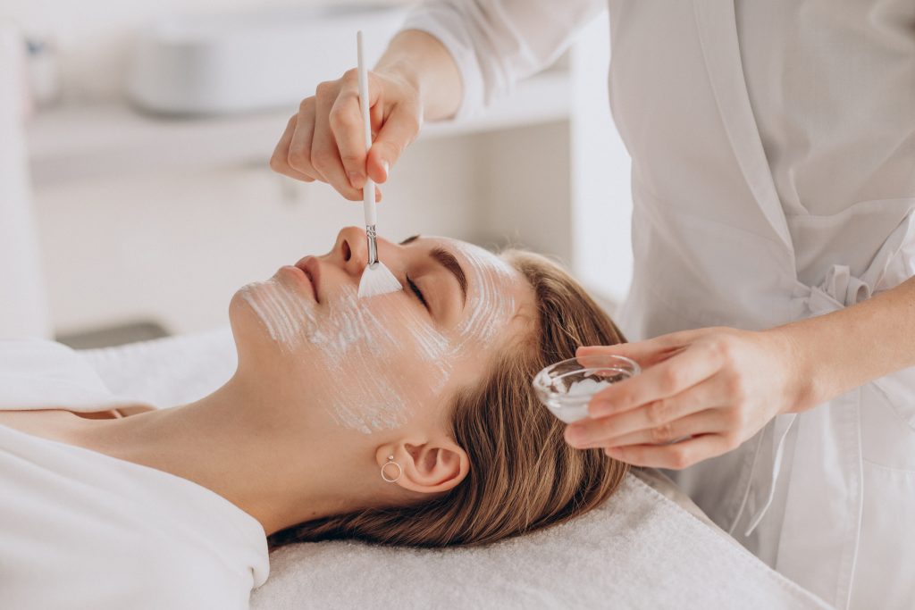 In questa foto una sposa mentre fa una maschera visto dall'estetista che la stende sul viso con un pennello a ventaglio