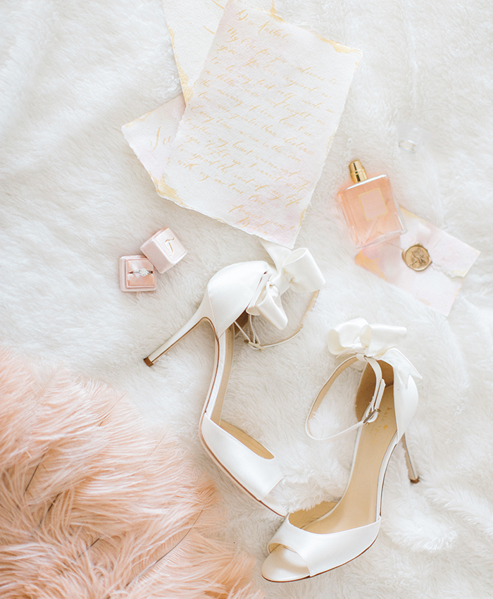 In questa foto scarpe da sposa open toe con cinturino alla caviglia e fiocco, un profumo, un anello con diamante e un partecipazione con ceralacca colore oro. Tutto è adagiato su un tappeto bianco