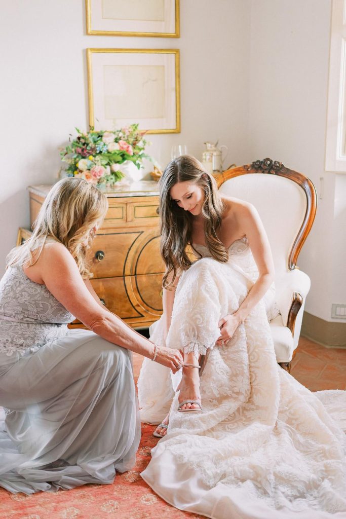 In questa foto una sposa seduta su una poltrona mentre la sua damigella chinata la aiuta a chiudere il cinturino dei suoi sandali gioiello