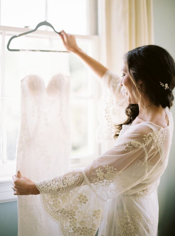 In questa foto una sposa in vestaglia di pizzo di tre quarti mentre guarda il suo abito da sposa a sirena davanti alla finestra 