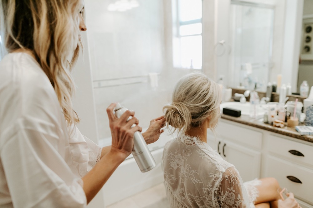 In questa foto della preparazione sposa un'amica le spruzza la lacca sulla sua acconciatura a chignon mentre lei sta seduta davanti allo specchio
