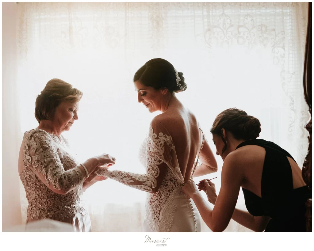 In questa foto la preparazione di una sposa mentre, davanti alla finestra della camera da letto, la mamma le chiude il bracciale sul polso e la sorella leggermente chinata le chiude il vestito sulla schiena 
