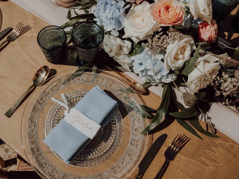 In questa foto una mise en place Country Chic con segnaposto matrimonio bianco con scritta calligrafica su tovagliolo celeste abbinato ai fiori del centrotavola. Il tovaglio è poggiato su un piatto di vetro trasparente che lascia intravedere la tavola di legno nudo