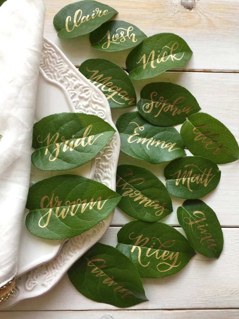 In questa foto foglie come segnaposto di nozze con nomi degli ospiti con scritta calligrafica colore oro