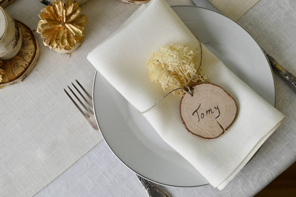 In questa foto un tronchetto di legno come segnaposto di nozze con il nome dell'ospite scritto a penna e legato ad un fiore bianco. Entrambi sono poggiati su un tovagliolo bianco 