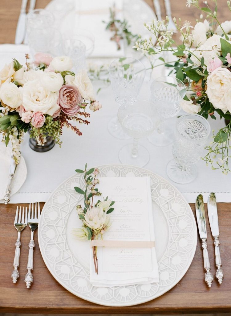 In questa foto un segnaposto matrimonio con fiore bianco e foglia poggiato su un menù matrimonio in una mise en place in stile rustic chic con tavola ricamata su tavola di legno nudo