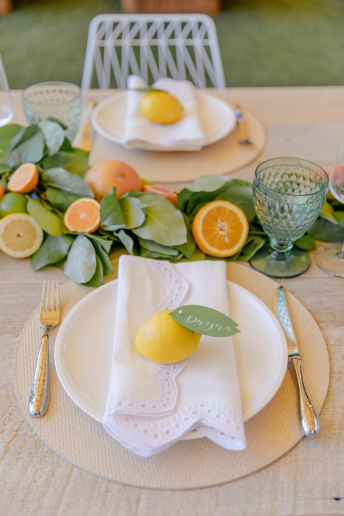 In questa foto segnaposto per matrimonio con limoni e nome dell'ospite scritto sulla foglia. Il limone è poggiato su un tovagliolo ricamato di colore bianco. La tavola è decorata con un runner di foglie e arance tagliate a metà