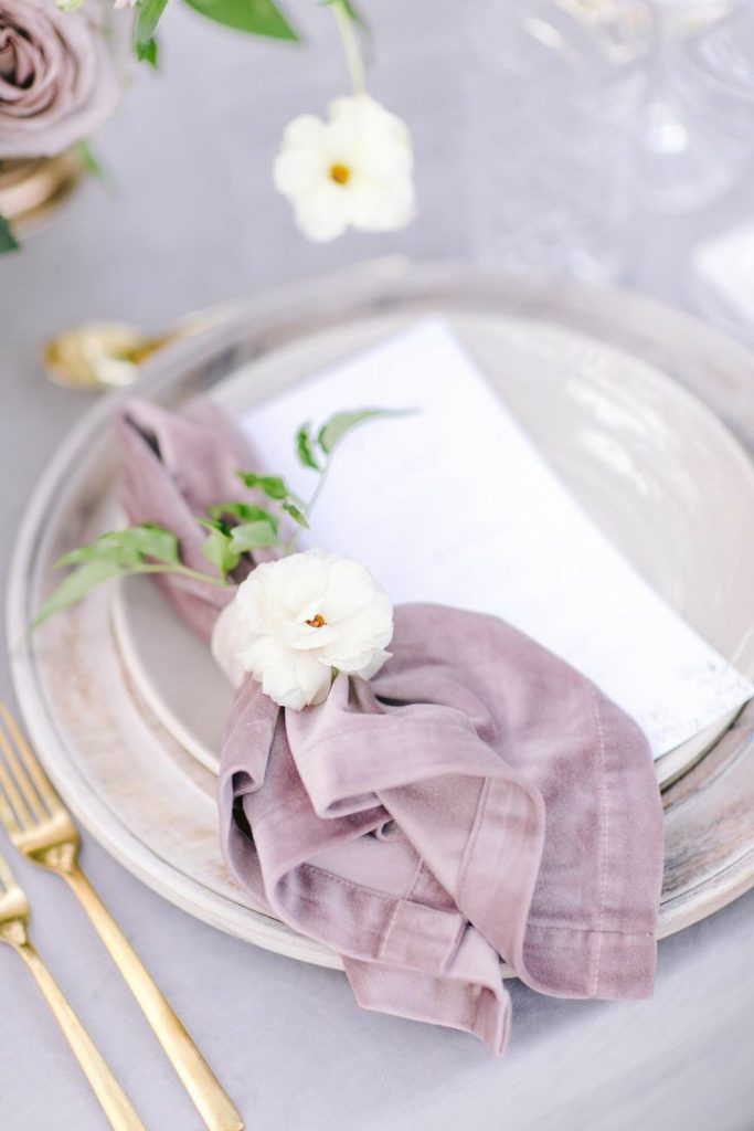 In questa foto un portatovagliolo lavanda e tovagliato in coordinato con un fiore bianco e posate oro