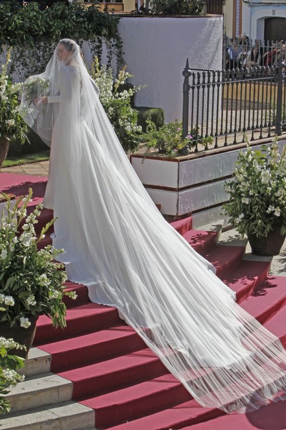 In questa foto la modella Eva Gonzalez avvolta dal suo velo da sposa Cattedrale nel giorno delle sue nozze ritratta su una scalinata con tappeto rosso circondata da fiori bianchi