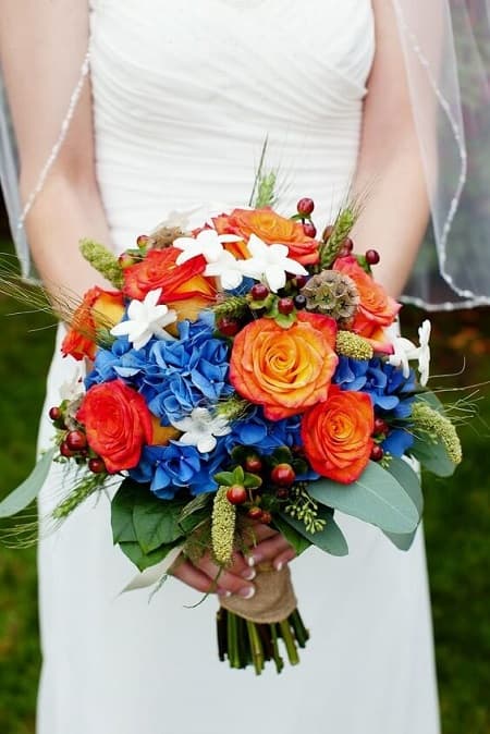 In questa foto, una sposa tiene in mano un bouquet rotondo realizzato con rose di colore arancione, ortensie blu, gelsomino bianco e bacche